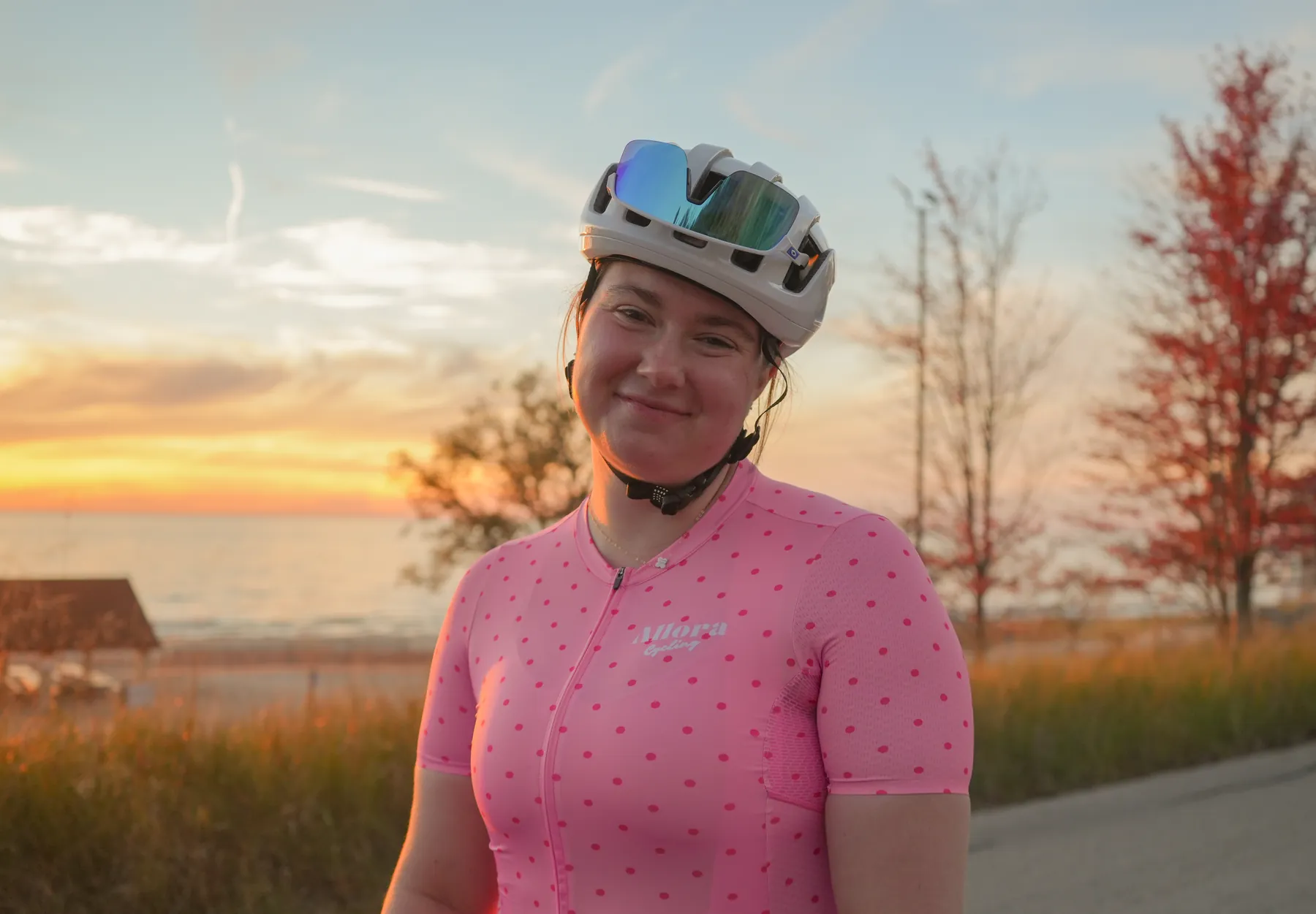 Addison smiling at the camera at sunset by the water, in her pink polka-dot Allora jersey with her helmet and sunglasses on