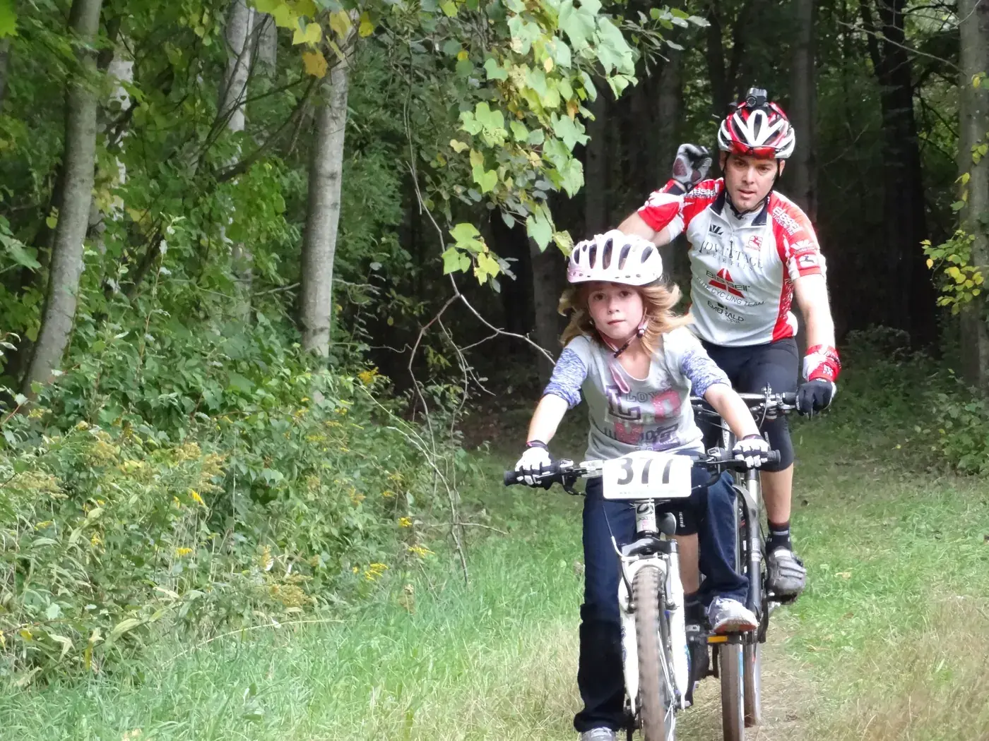 Young Addison racing her mountain bike down a wooded trail, an adult rider close behind her